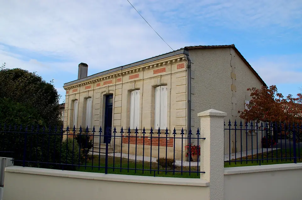 Échoppe girondine en pierre de taille soulignée de briques, avec hautes fenêtres à volets clairs derrière une grille et un muret.