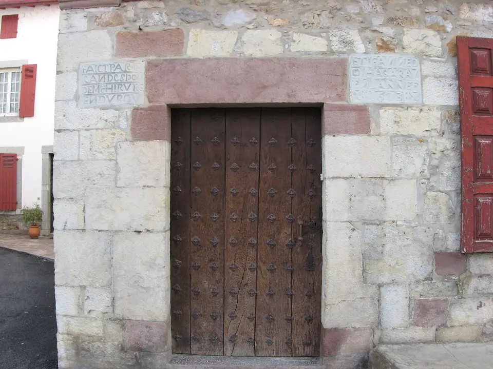 Porte d'entrée en bois cloutée dans un encadrement de grès rose, sur une maison basque aux volets rouges.